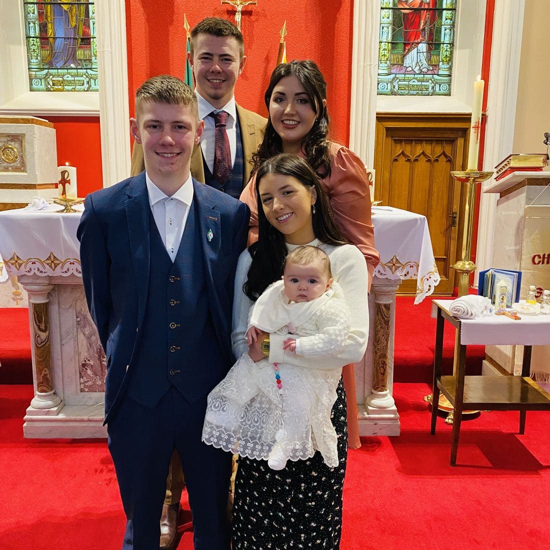 Córa in a long white christening gown, held by Erin at the altar of St Benignus Church with Conor and family gathered around