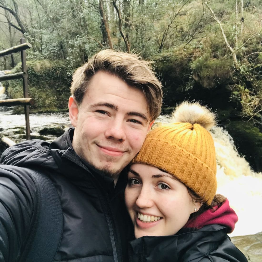 Conor and Erin cheek to cheek in winter coats and a mustard bobble hat, a waterfall running through the woods behind them