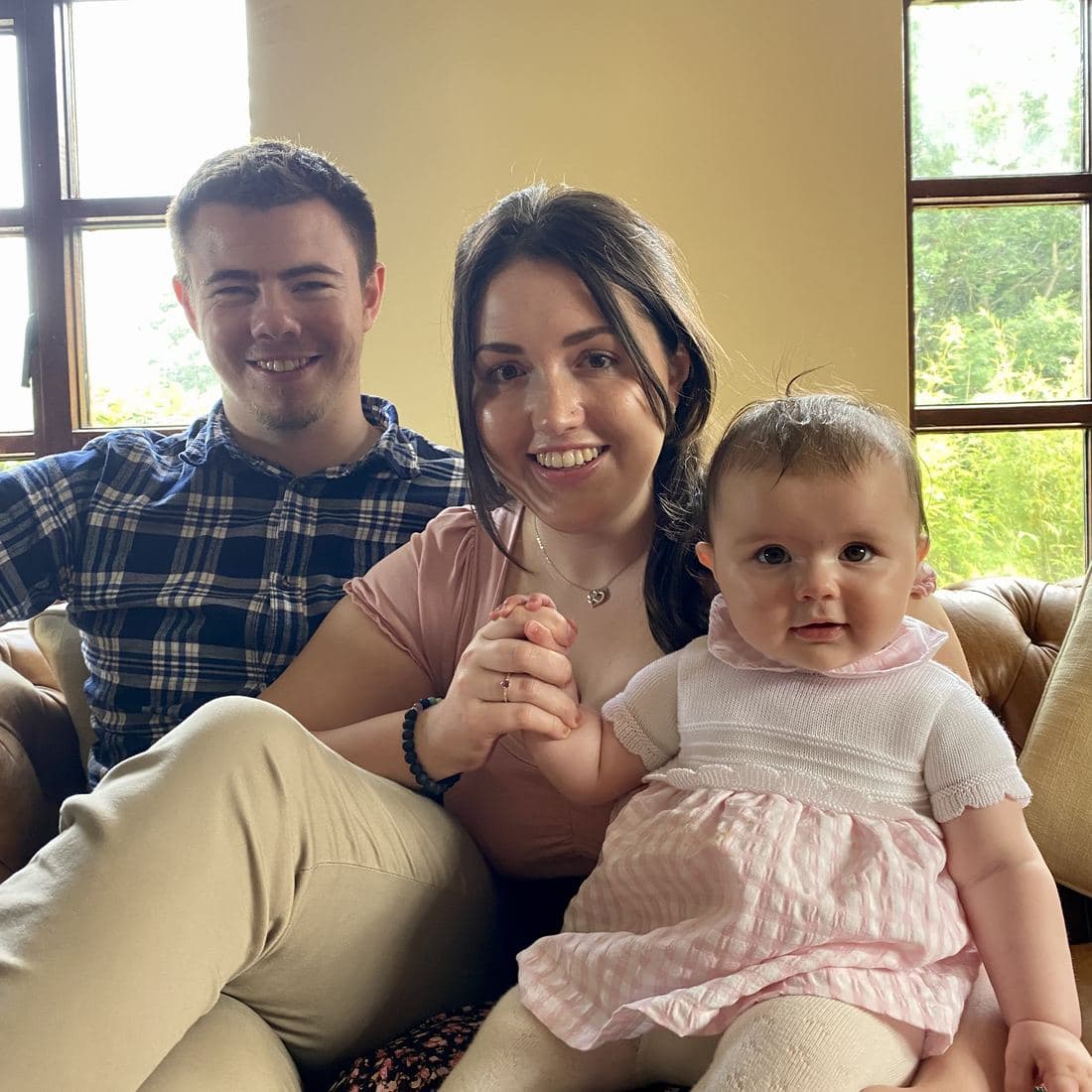 Conor and Erin sitting together on a sofa in summer light with Córa on Erin’s lap, holding her hand